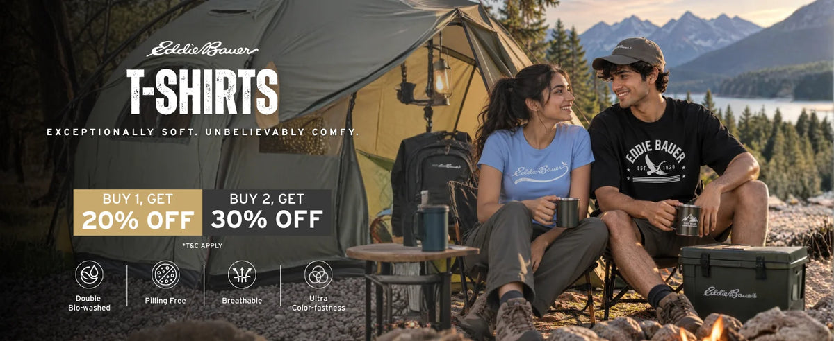 A promotional outdoor scene showing a man and woman sitting by a campfire in front of a tent, wearing Eddie Bauer T-shirts. Large text on the left reads 'Eddie Bauer T-Shirts' with sale copy, and the background shows mountains, trees, and a lake.