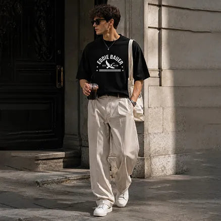 A man wearing a black graphic t-shirt with 'Eddie Bauer' printed in white, paired with beige wide-leg trousers, white sneakers, and a white shoulder bag, walking outdoors against a stone building.