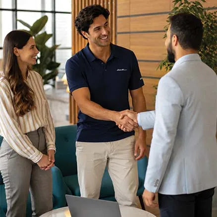A man in a navy blue polo shirt with a small white script logo on the chest shakes hands with another man in a light blazer, while a woman in a striped blouse stands beside them in a modern office lobby.