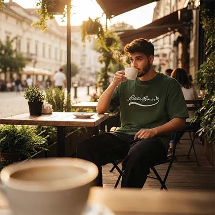 A man sitting at an outdoor cafe table in warm golden hour light, wearing a dark green t-shirt with a white 'Eddie Bauer' script logo across the chest.