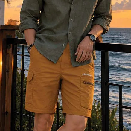 A man wearing mustard yellow cargo shorts and an olive green button-up shirt, standing on a balcony overlooking the ocean at sunset.