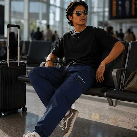 A man wearing a black t-shirt and navy blue cargo pants sits on a airport waiting area bench next to a black suitcase.