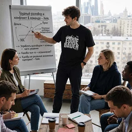 A man in a black t-shirt with a white 'EDDIE BAUER' logo stands next to a whiteboard during a business meeting, with several colleagues seated around him in a modern office setting with a city skyline background.