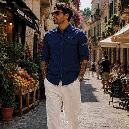 A man wearing a dark blue button-down shirt with 'Johnnie Deppa' and 'UNIQ' branding, paired with off-white trousers, walking down a sunny, pedestrian-friendly European street lined with shops and cafes.