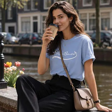 A woman sitting outdoors by a canal wearing a light blue graphic T-shirt with 'Elles & Bauer' printed on the chest, paired with black pants and a beige shoulder bag.