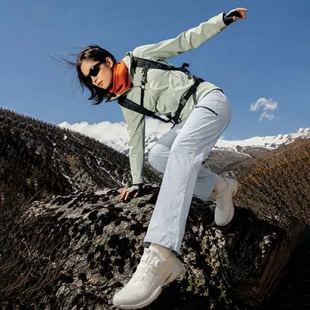 A woman in outdoor gear, including a light sage green jacket, white pants, white hiking boots, and an orange neck gaiter, is posed dynamically on a rocky mountain peak under a clear blue sky.