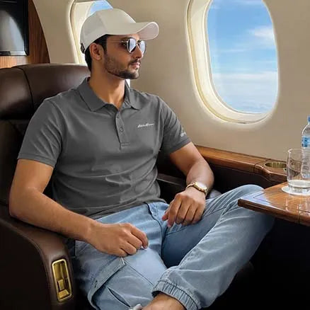 A man wearing a gray polo shirt, light blue jeans, a white cap, and sunglasses sits in a luxurious airplane business class seat with a window view.