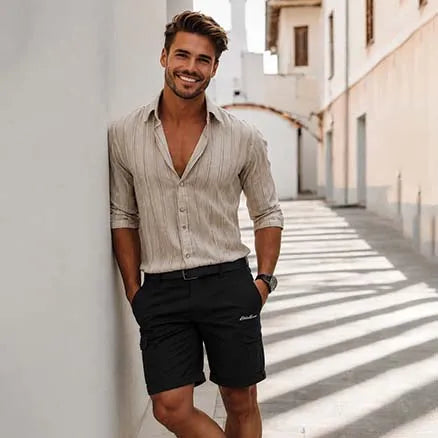 A smiling man stands in a sunlit outdoor alley wearing a light beige vertically striped button-down shirt with rolled sleeves and black cargo shorts.