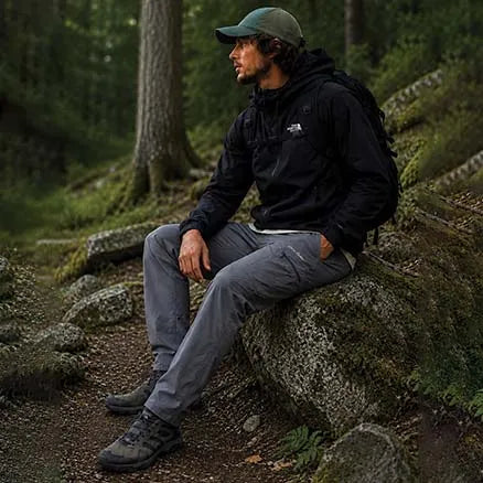 A man wearing a black The North Face jacket, gray hiking pants, and a green cap sits on a mossy rock in a forest setting.
