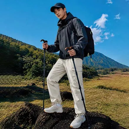 A man hiking outdoors in a mountain landscape, wearing a dark gray hooded jacket, light beige cargo pants, white sneakers, a black backpack, and holding two black trekking poles.