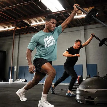 A man in a teal Fossil Bauer t-shirt and black shorts exercises with a barbell in a gym, with another man working out in the background.