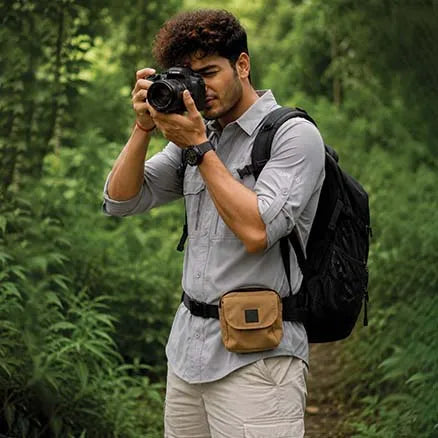 A man in a forest setting wearing a light gray button-up shirt, beige pants, a black backpack, and a tan waist pouch bag, holding a DSLR camera.