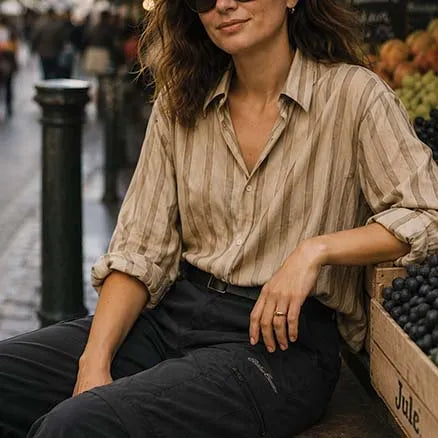 A woman wearing a beige and brown vertically striped button-down shirt and dark trousers sits next to a crate of blueberries in an outdoor market setting.