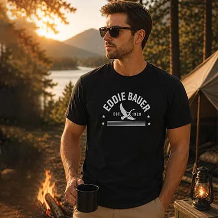 A man wearing a black graphic t-shirt with a white 'Eddie Bauer Est. 1920' logo, standing outdoors at a campsite with a campfire, tent, and mountains in the background.