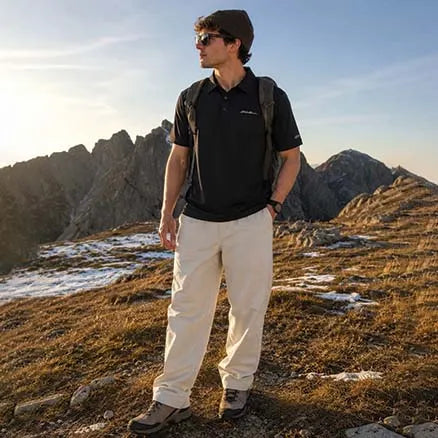 A man wearing a black short-sleeve polo shirt, beige hiking pants, and a dark backpack stands on a mountain ridge during golden hour, looking off-camera. The background features rugged rocky mountains under a clear blue sky.