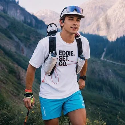 A man trail running in a mountainous outdoor setting, wearing a white t-shirt with the text 'EDDIE. SET.