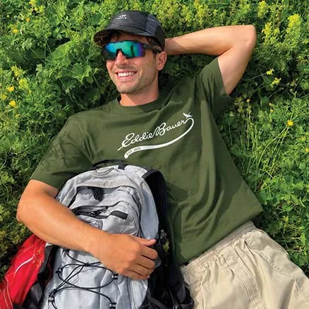 A man lying in green grass outdoors, wearing an olive green Eddie Bauer t-shirt with a white script logo, a black cap, and sunglasses, and holding a gray hiking backpack.