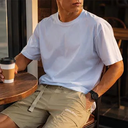 A man sitting at a cafe table wearing a light blue crew-neck t-shirt and khaki cargo shorts, holding a paper coffee cup.