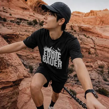 A man wearing a black t-shirt with a white 'EDDIE BAUER' graphic, black shorts, and a black cap, posing in a red rock desert canyon while climbing a chain.