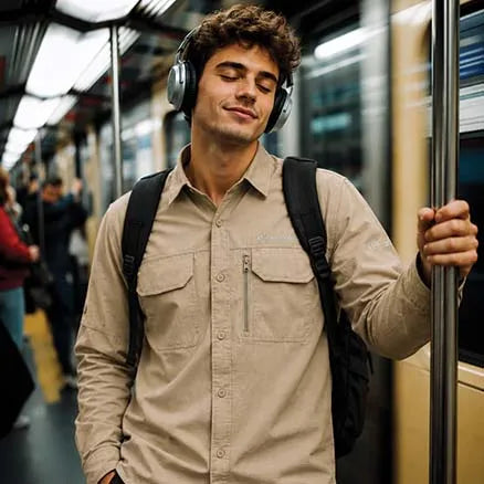 A man with curly brown hair wearing a beige button-up shirt, black backpack, and silver over-ear headphones stands on a subway train holding a metal pole.