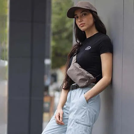 A woman wearing a black short-sleeve t-shirt with a small white logo on the chest, light blue cargo pants, a taupe cap, and a taupe crossbody bag, leaning against a gray wall.