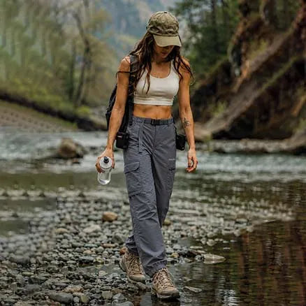 A woman hiking through a shallow river with rocks, wearing a beige crop top, gray cargo pants, brown hiking boots, and a camouflage cap, carrying a water bottle and a backpack.