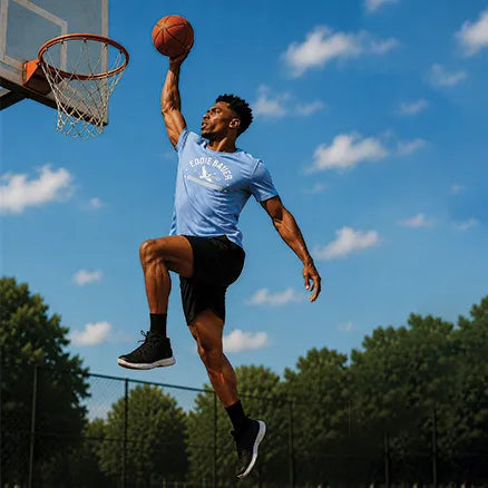 A man in a light blue 'LOONEY TUNES' t-shirt and black shorts performs a dynamic basketball dunk in mid-air on an outdoor court under a blue sky with clouds.