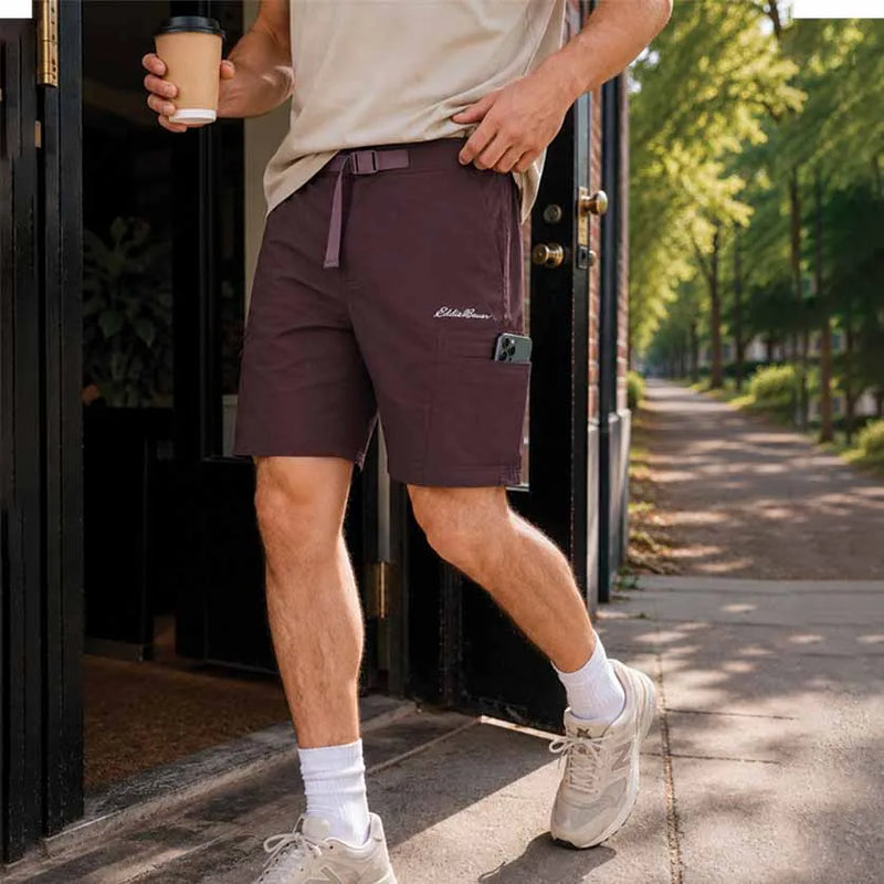 A man wearing dark plum cargo shorts, a beige t-shirt, white socks, and beige sneakers walking out of a doorway on a sunny day.