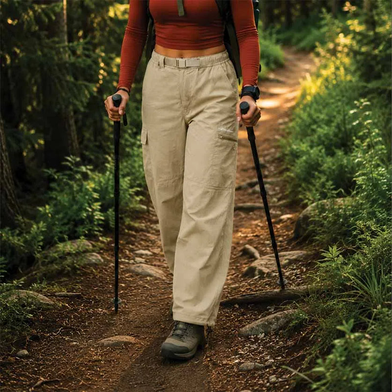 A person hiking on a forest trail wearing beige cargo pants, a rust-orange long-sleeve crop top, and hiking boots, holding black trekking poles.