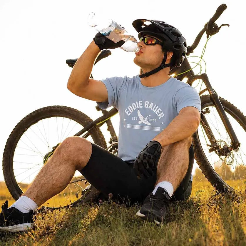 A man wearing a light blue graphic t-shirt with 'EDDIE BAUER EST. 2012' sits on a grassy hill behind a bicycle, drinking from a water bottle.