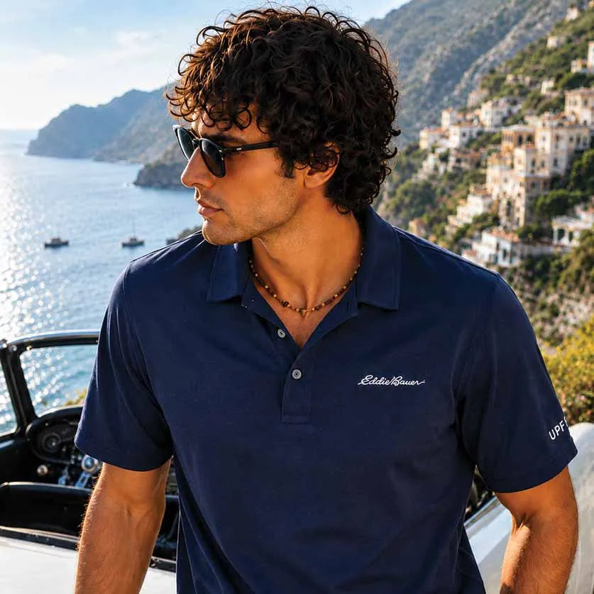 A man wearing a navy blue Eddie Bauer polo shirt and sunglasses, standing outdoors near a boat with a scenic coastal town and sea in the background.