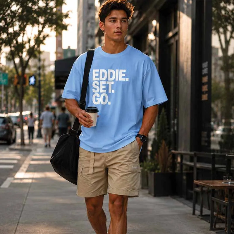 A man walking on a city sidewalk wearing a light blue t-shirt with the text 'EDDIE. SET.
