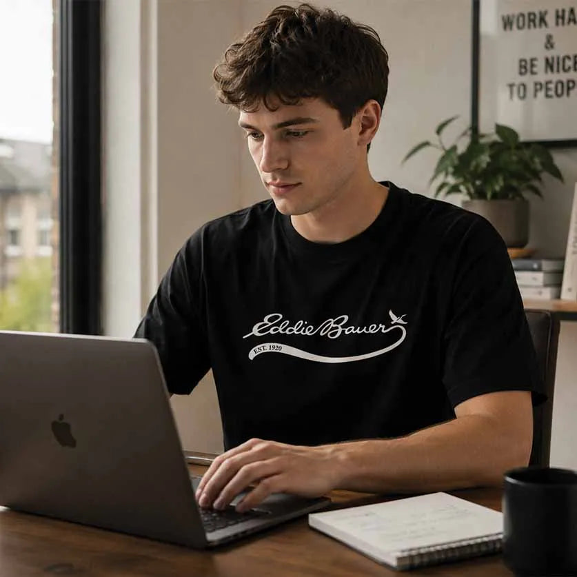 A man wearing a black crewneck t-shirt with a white 'Eddie Bauer' logo sits at a wooden desk using a laptop in a bright room.