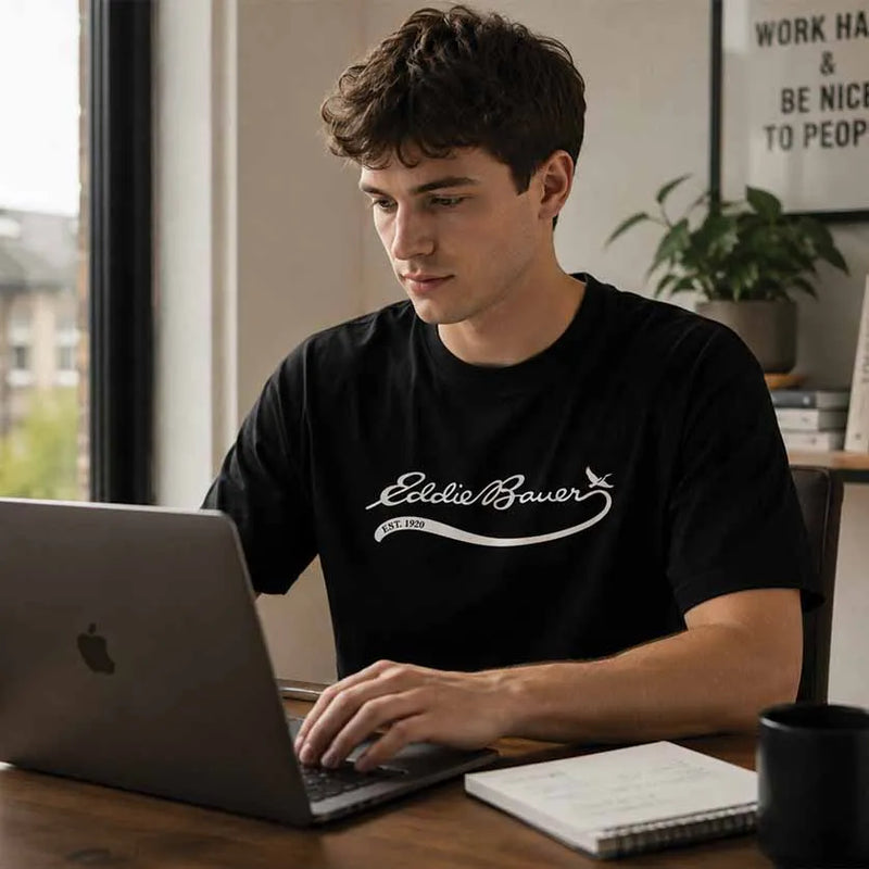 A man wearing a black crewneck t-shirt with a white 'Eddie Bauer' logo sits at a wooden desk using a laptop in a bright room.