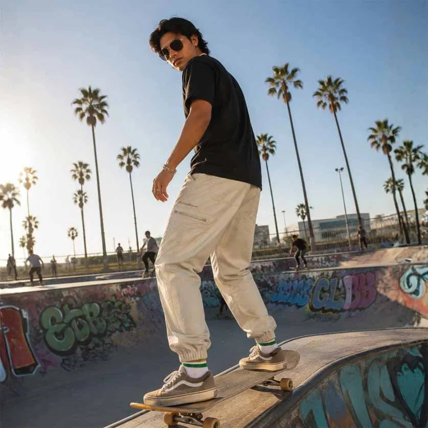 A man wearing a black t-shirt, beige cargo pants, and beige sneakers stands on a skateboard at a graffiti-covered skatepark with palm trees in the background.
