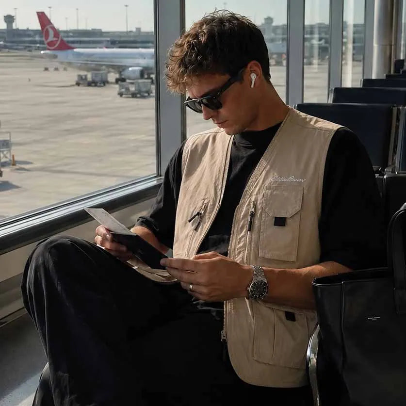 A man sitting in an airport lounge by a window, wearing a beige utility vest over a black t-shirt and black pants, looking at his phone. A Turkish Airlines plane is visible outside the window.