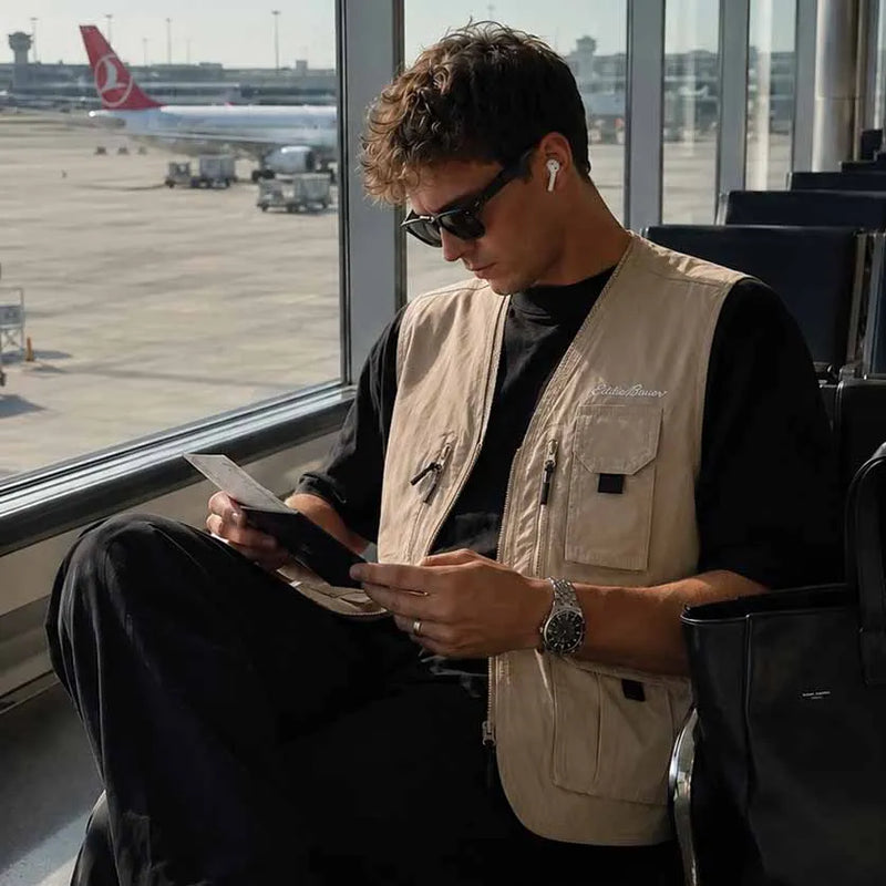 A man sitting in an airport lounge by a window, wearing a beige utility vest over a black t-shirt and black pants, looking at his phone. A Turkish Airlines plane is visible outside the window.