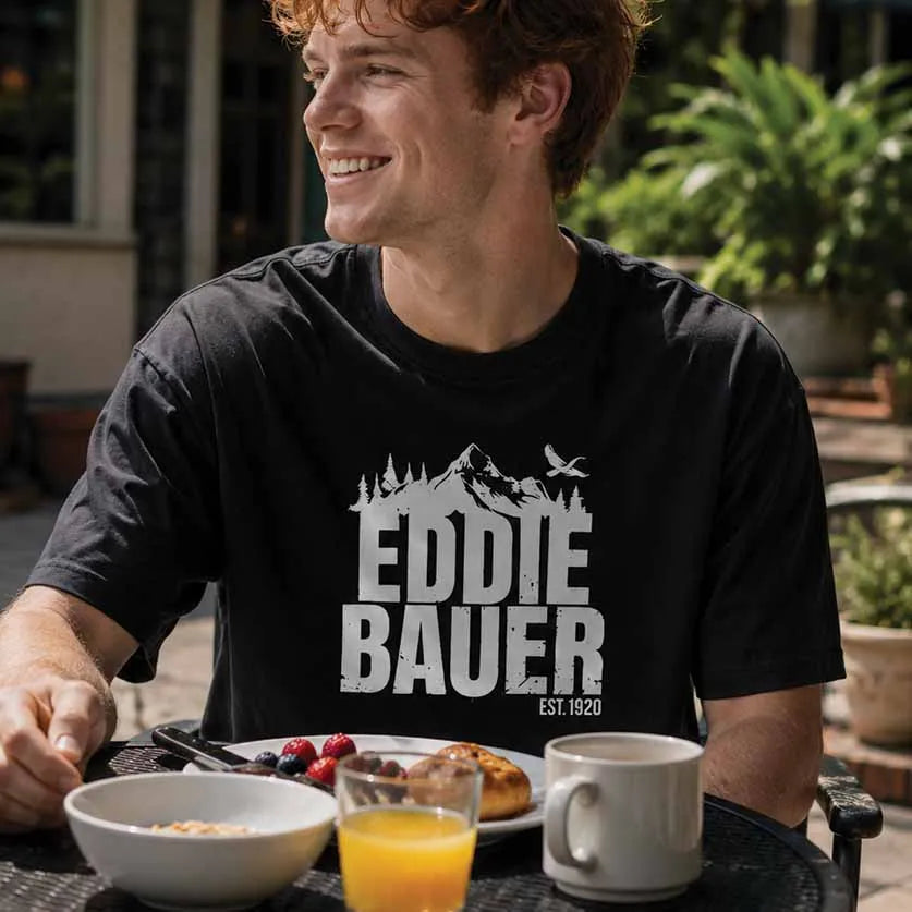 A smiling man sitting at an outdoor patio table wearing a black graphic t-shirt with a white distressed mountain and bird logo and the text 'EDDIE BAUER EST. 1920'.