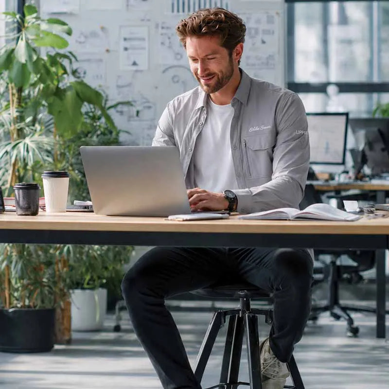 A man sits on a stool at a wooden desk in a bright office, using a silver laptop. He is wearing a light gray button-up shirt over a white t-shirt and dark pants.
