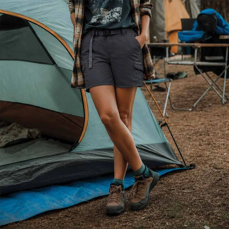 A person standing outdoors beside a tent at a campsite, wearing dark gray hiking shorts, brown hiking boots, teal socks, and an open plaid shirt over a dark top.
