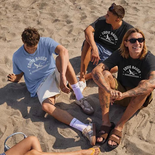 Three men sitting on a sandy beach wearing matching Eddie Bauer t-shirts. One man wears a light blue t-shirt, while the other two wear black t-shirts.