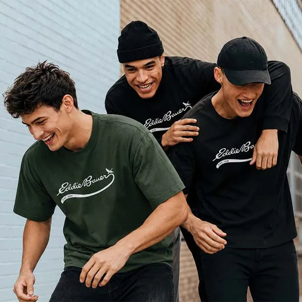 Three men laughing outdoors while wearing Eddie Bauer branded t-shirts in dark green, black, and dark gray. One man wears a black beanie, and two wear black caps.