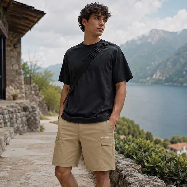 A man wearing a black short-sleeve t-shirt and beige cargo shorts stands outdoors on a stone path with a scenic lake and mountains in the background.