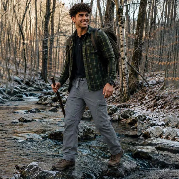 A man hiking through a rocky stream in a forest, wearing a green and black plaid flannel shirt over a black polo, gray cargo pants, and brown hiking boots, with a backpack on.