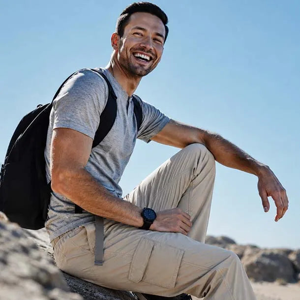 A smiling man wearing a gray t-shirt, beige cargo pants, and a black backpack sits outdoors on a rocky surface under a clear blue sky.