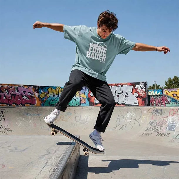 A young man skateboarding in a concrete bowl at a graffiti-covered outdoor skatepark, wearing a light sage green t-shirt with 'EDDIE BAUER' printed in white on the front, black pants, and white sneakers.