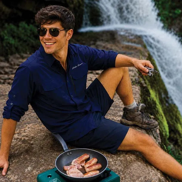 A man wearing a navy blue long-sleeve shirt and matching shorts sits on a rock in front of a waterfall, cooking sausages in a pan on a portable stove.