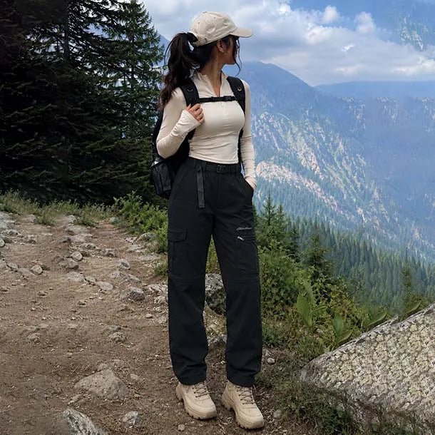 A woman stands on a mountain trail wearing a beige long-sleeve top, black cargo pants, beige hiking boots, and a beige baseball cap, with a black backpack against a backdrop of green pine trees and blue mountains.