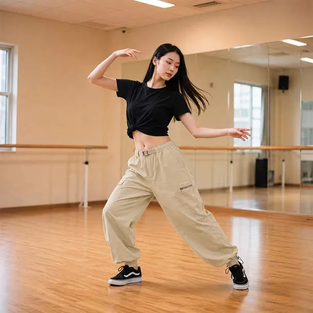 A woman posing in a dance studio, wearing a black short-sleeve crop top and beige cargo pants with black sneakers.