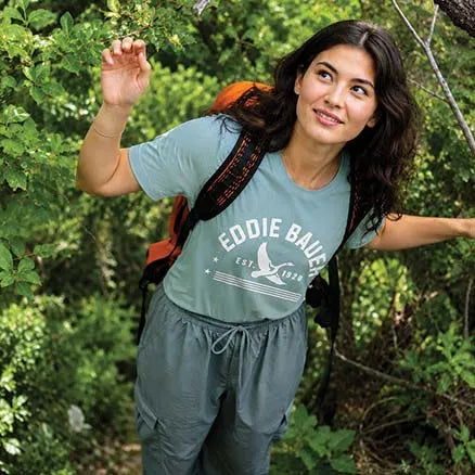 A woman hiking outdoors in a light blue-green graphic t-shirt with 'EDDIE BAUER EST. 1954' printed on the front, paired with matching drawstring cargo pants and a backpack.